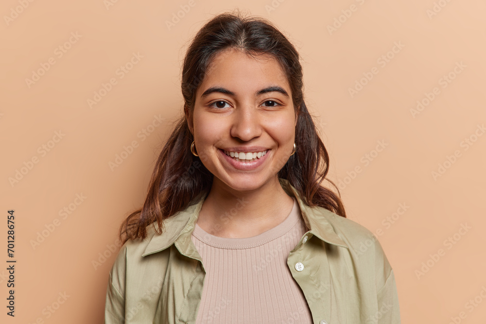 Portrait of lovely dark haired Iranian woman smiles pleasantly has glad expression dressed in casual shirt looks directly at camera isolated over brown background. Positive human emotions concept Portrait of lovely dark haired Iranian woman smiles pleasantly has glad expression dressed in casual shirt looks directly at camera isolated over brown background. Positive human emotions concept