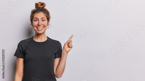 Horizontal shot of pretty young European woman with hair bun smiles pleasantly dressed in casual black tshirt points index finger on empty space isolated over white background. Look at this. photo