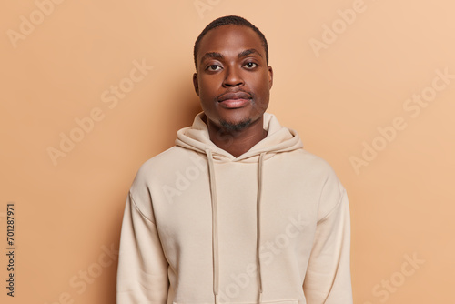 Horizontal shot of handsome dark skinned African man with small beard full lips big eyes looks directly at camera dressed in casual sweatshirt poses against brown background. Human face expressions