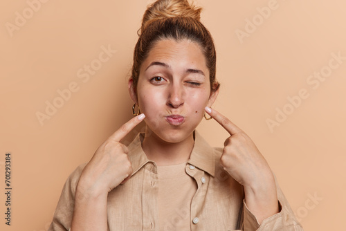 Photo of lovely young woman with hair bun points index finger at cheeks holds breath makes funny grimace wears shirt isolated over brown background tries to make someone laughing. Hey look at me photo