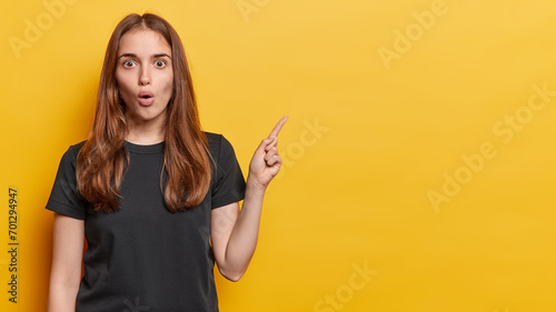 Omg what cool offer. Waist up shot of amazed long haired woman stands speechless has widely opened eyes and mouth wears casual black t shirt isolated over yellow background points at blank dpace photo
