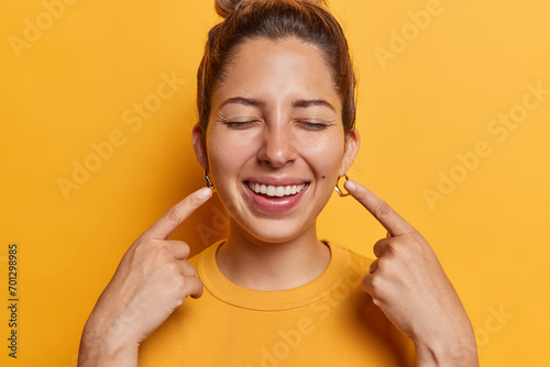Positive overjoyed young European woman with healthy glowing skin points at her broad smile shows perfect white teeth after whitening procedures dressed casually isolated over yellow background