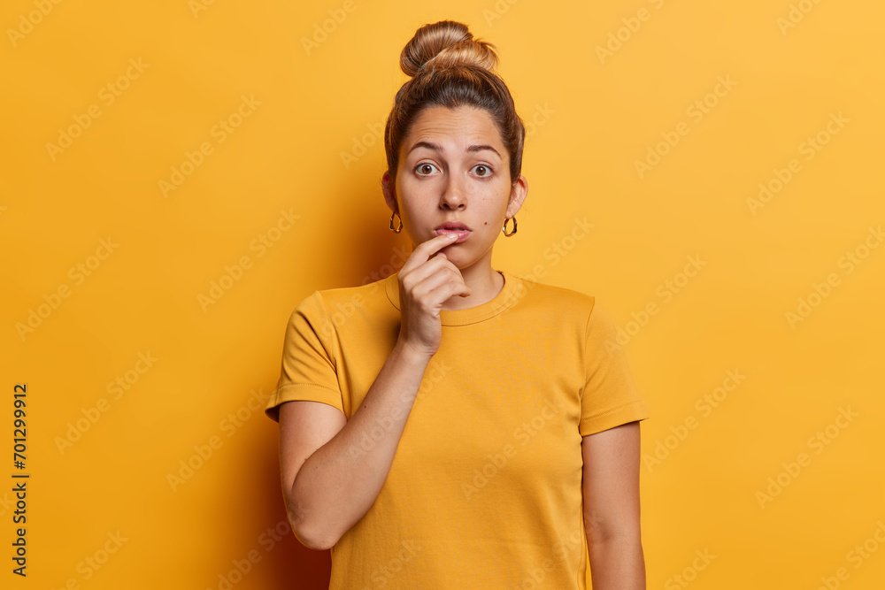 Horizontal shot of shocked lovely woman with hair bun stares impressed at camera keeps finger on lips dressed in casual t shirt isolated over vivid yellow background reacts to something impressive Horizontal shot of shocked lovely woman with hair bun stares impressed at camera keeps finger on lips dressed in casual t shirt isolated over vivid yellow background reacts to something impressive