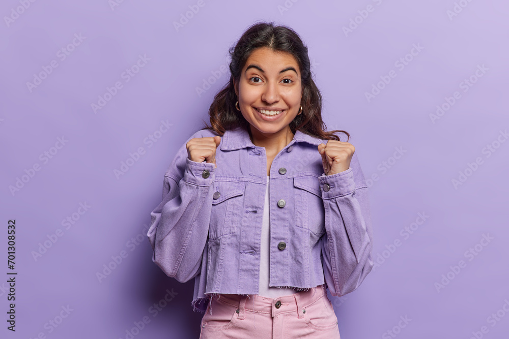 Photo of dark haired Iranian clenches fists cheers over triumph celebrates good news smiles toothily wears jacket and pink trousers isolated over purple background. Success and celebration concept