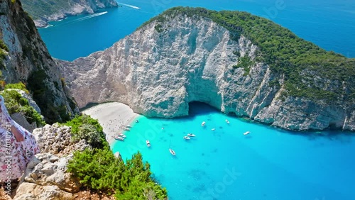 Aerial view of a beautiful woman looking at Navagio Pebble Beach with a shipwreck and turquoise water. Female tourists enjoying summer vacation at the cove reached by boats in Zakynthos, Greece. photo