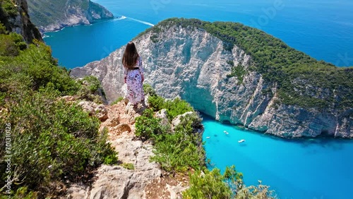 Aerial view of a beautiful woman looking at Navagio Pebble Beach with a shipwreck and turquoise water. Female tourists enjoying summer vacation at the cove reached by boats in Zakynthos, Greece.