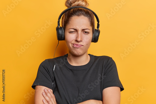 People emotions concept. Indoor photo of young European girl standing in centre isolated on yellow background with crossed raised hands listening to music on headphones dissapointed to hear new song photo