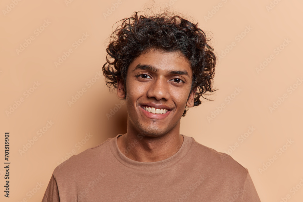 Portrait of merry positive curly haired Hindu man smiles pleasantly being in good mood dressed in casual t shirt looks directly at camera isolated over brown background. People and emotions concept Portrait of merry positive curly haired Hindu man smiles pleasantly being in good mood dressed in casual t shirt looks directly at camera isolated over brown background. People and emotions concept