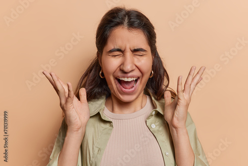 Photo of emotional brunette attractive Iranian girl exclaims loudly keeps palms raised up and reacts to something dressed in casual shirt isolated over brown background. Human reactions concept
