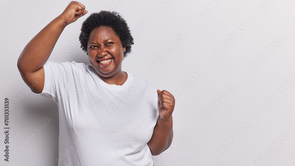 Photo of overjoyed dark skinned plump woman makes winner gesture raises arms and clenches fists feels like winner dressed in casual t shirt isolated over white background copy space for your advert - obrazy, fototapety, plakaty 