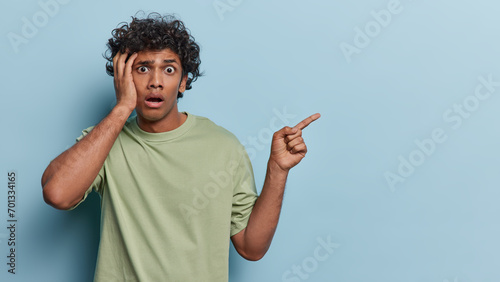 People emotions concept. Studio waist up of young surprised Hindu male standing on left isolated on blue background pointing at blank space for your promotion wearing olive t shirt looking straight photo