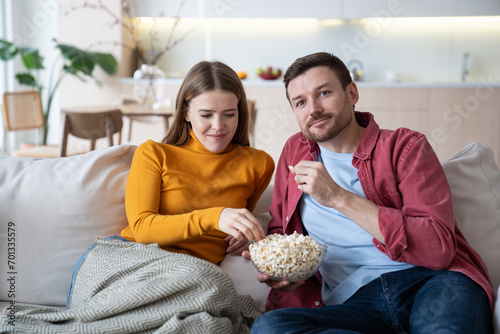 Happy married couple spending weekends at home, sitting on couch, watching TV. Smiling cheerful wife eats popcorn, intrigued husband watches favourite show, video, interesting film episode on TV