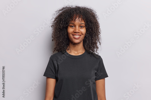 Waist up shot of pretty curly haired woman smiles happily dressed in casual black t shirt keeps arms down looks gladfully at camera isolated over white background. People and emotions concept © Wayhome Studio