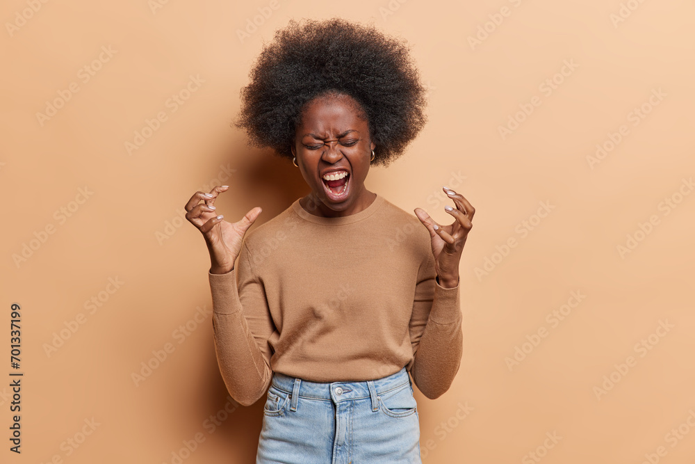 Annoyed outraged dark skinned young African woman keeps hands in furious gesture screams loudly being irritated because of something wears casual jumper and jeans isolated over brown background. Annoyed outraged dark skinned young African woman keeps hands in furious gesture screams loudly being irritated because of something wears casual jumper and jeans isolated over brown background.