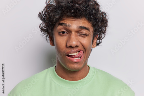 Human facial expressions and fun concept. Funny curly haired Hindu man winks eye and sticks out tongue tries to make you laugh dressed in casual green t shirt isolated over white background. photo