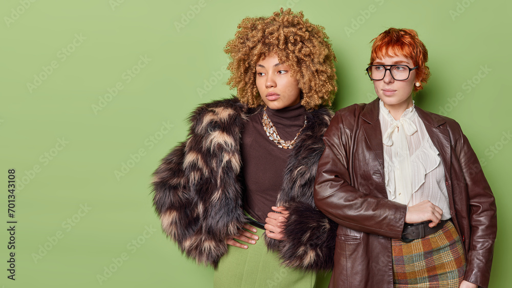 Horizontal shot of two pensive women being deep in thoughts stand closely to each other dressed in fashionable clothing isolated over green background with copy space for your advertisement.