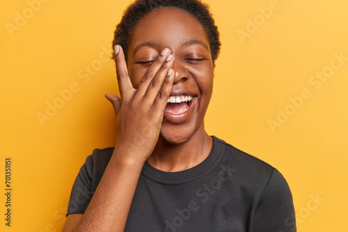 People positive emotions concept. Studio photo of young happy smiling African american lady with closed eyes standing in centre isolated on yellow background covering face with palm wearing t shirt