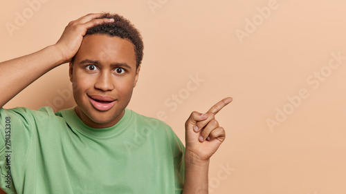 People emotions concept. Studio shot of young confused African american man holding hand on head not knowing what to do pointing at blank space for promotion standing on left on beige background