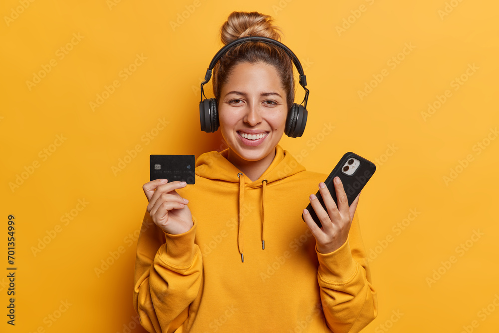 Fototapeta premium Positive emotions concept. Indoor waist up of young glad smiling European lady listening to music on headphones standing isolated on yellow background holding smartphone and card wearing hoodie
