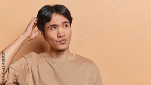Horizontal shot of confused young Japanese man scratches head and concentrated with hesitation asie thinks about something makes decision dressed in casual t shirt isolated over brown backgroud