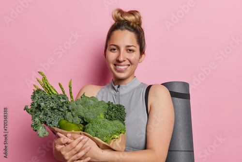 Balanced and nourishing lifestyle concept. Positive fit woman with hair gathered in bun holds paper bag full of fresh green vegetables rubber fitness mat smiles happily isolated over pink background