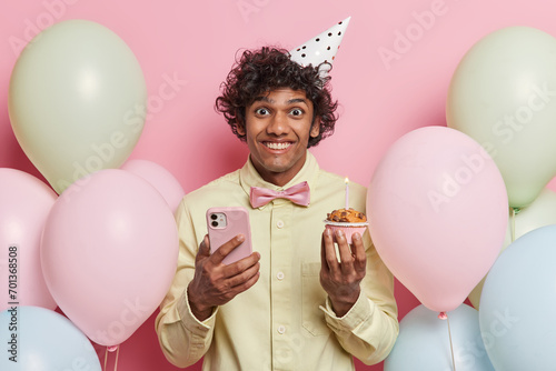 Positive young Hindu man with curly hair smile pleasantly holds mobile phone and cupcake celebrates birthday wears cone hat yellow shirt and bowtie surrounded by inflated balloons poses indoor © wayhome.studio 