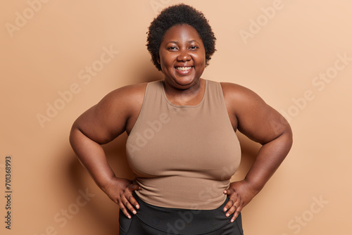 Waist up shot of cheerful African woman with curly hair keeps hands on waist smiles gladfully being body positive wears t shirt isolated over brown background. Plus size female model stands indoor