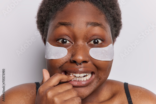 Beauty and cosmetology concept. Indoor shot of young scared plump African american woman applying white patches biting finger wearing black undershirt looking straight at camera standing isolated
