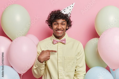 Positive curly haired Hindu man points at himself smiles pleasantly asks who me wears cone hat and yellow shirt has festive mood poses among inflated colorful balloons celebrates special occasion © wayhome.studio 