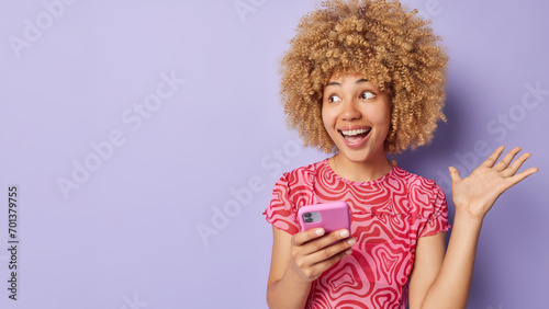People positive emotions concept. Indoor photo of young pretty cheerful smiling African american lady standing on right isolated on purple background holding smartphone with blank space for promotion