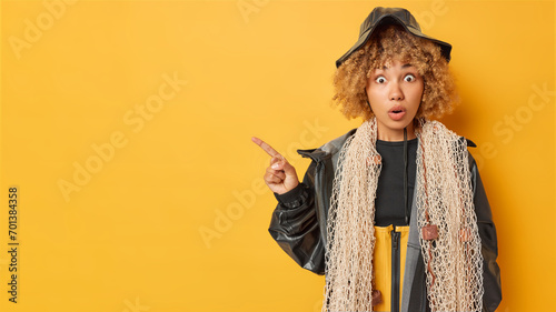 Indoor shot of impressed fisherwoman showcases her expertise poses with net around neck has fishing expedition wears black hat and leather jacket points index finger on empty space for your text photo