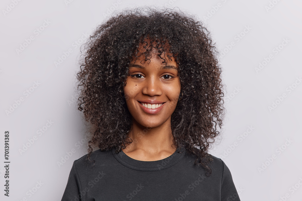 Portrait of cheerful curly haired teenage girl with healthy skin perfect teeth dressed in casual black t shirt looks directly at camera isolated over white background. People and positive emotions - obrazy, fototapety, plakaty 