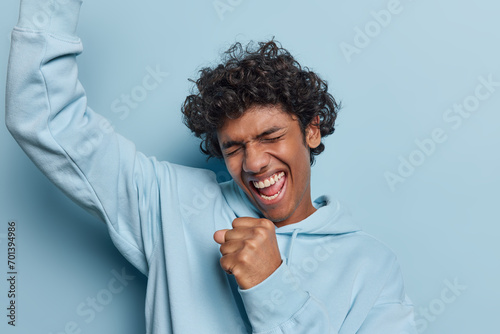 Handsome young Hindu man with dark curly hair keeps fist near mouth as if microphone dances carefree wears casual hoodie isolated over blue background enjoys favorite music catches every bit