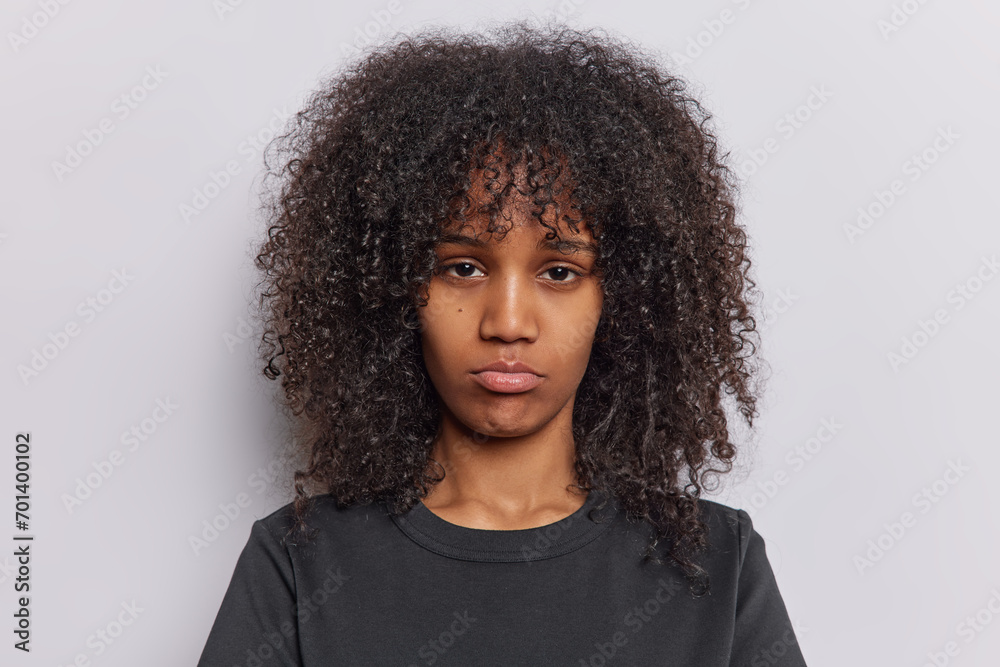 Portrait of upset curly haired woman purses lips has disappointed face being discontent by something wears black t shirt isolated over white background. People and negative emotions concept. - obrazy, fototapety, plakaty 