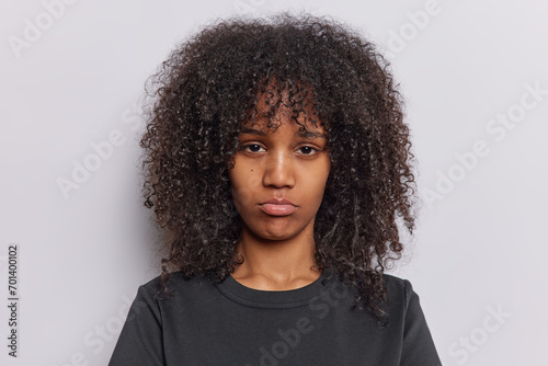 Portrait of upset curly haired woman purses lips has disappointed face being discontent by something wears black t shirt isolated over white background. People and negative emotions concept.