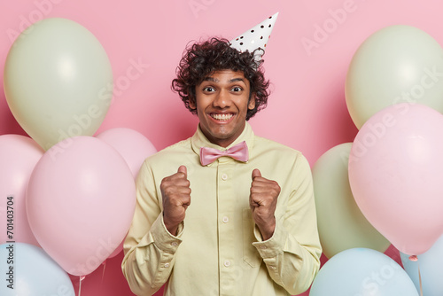 Positive young Hindu man with dark curly hair clenches fists celebrates success gets congratulations on birthday surrounded by inflated colorful balloons expresses happy emotions. Special occasion © Wayhome Studio