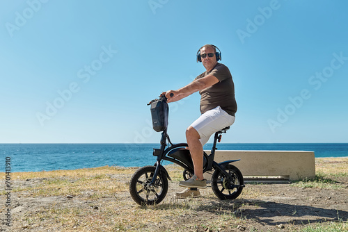 Overweight middle-aged man with headphones riding on electrical bicycle on the beach. Mature man in wireless headphones enjoying freedom in seaside.