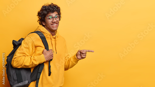 Horizontal shot of handsome curly haired Hindu man wears transparent eyeglasses and casual hoodie poses with backpack on shoulder indicates index finger on copy space isolated over yellow background © wayhome.studio 