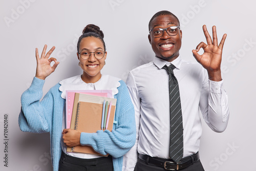 Schooling. Indoor waist up of young happy smiling broadly African american guy and Latin lady standing in centre on white background making ok signs holding books and notebooks in smart clothes