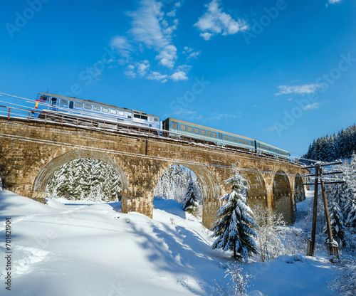 Stone viaduct (arch bridge) on railway through mountain snowy fir forest. Snow drifts on wayside and hoarfrost on trees and electric line wires.