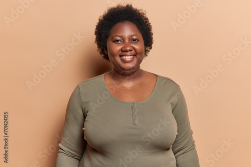 Portrait of cheerful plump adult African woman with short dark curly hair smiles pleasantly being in good mood happy to have pleasant conversation dressed casually isolated over brown background.