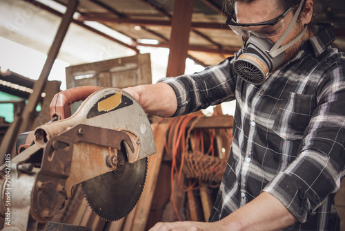 Male carpenter wearing protective mask using electric circular saw cutting wood board at workshop studio photo