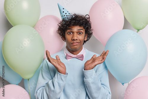 Birthday celebration concept. Indoor photo of young confused Hindu having party guy standing in centre on background of colourful balloons wearing festive clothes spreading palms with hesitation © wayhome.studio 