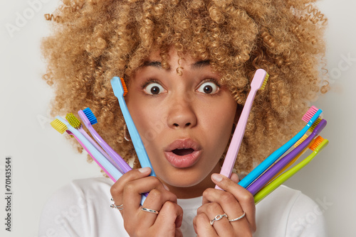 Headshot of curly haired speechless woman with blonde curly hair holds colorful toothbrushes chooses best one to clean teeth stares with omg expression at camera poses against white background. photo