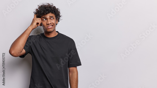 Waist up shot of confused curly haired young Hindu man scratches head and looks pensively aside thinks about something wears casual black t shirt isolated over white background. Hmm let me think