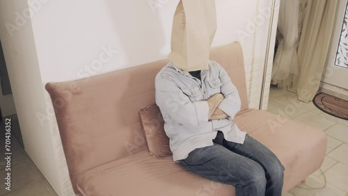 A man wearing a breadbag face, sitting on the sofa at home, no expression on his paper mask; doing nothing, feeling bored.
 photo