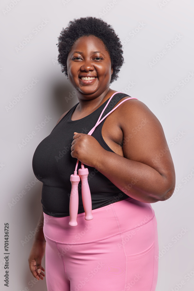 Sport and training concept. Positive overweight dark skinned adult woman dressed in activewear exercising with jumping rope dressed in black t shirt and leggings poses against white background.