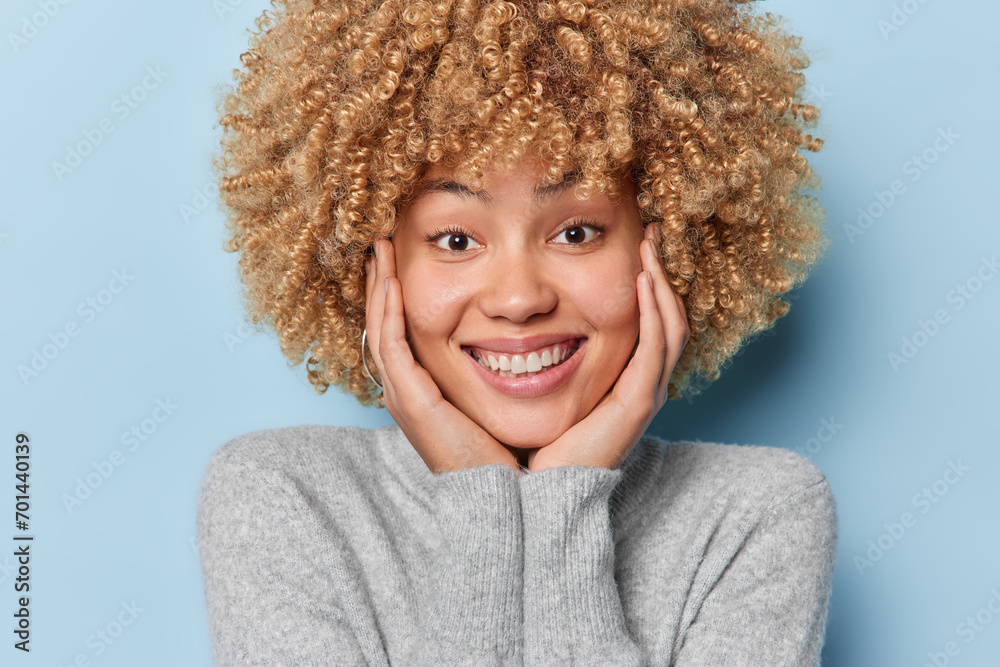 Portrait of pretty young woman with blonde curly hair keeps hands on cheeks smiles tenderly focused at camera dressed in casual grey jumper isolated over blue background. Positive emotions concept Portrait of pretty young woman with blonde curly hair keeps hands on cheeks smiles tenderly focused at camera dressed in casual grey jumper isolated over blue background. Positive emotions concept