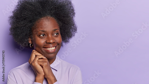 Studio shot of pretty dark skinned lady with bushy curly hair keeps hands near face concentratedd aside has gentle smile dreamy expression dressed formally isolated over purple background copy space photo