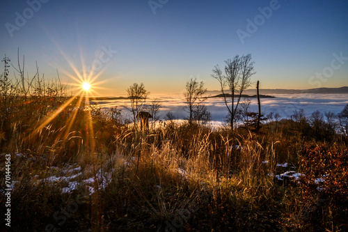Sunset over the winter landscape. Inverse weather with fog in the valleys. The sun with sun rays in the blue sky. In the foreground trees in winter without leaves.